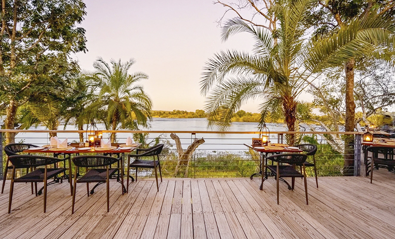 Terraza de madera de Baines Restaurant, con mesas iluminadas por faroles y palmeras frente al Zambeze al atardecer