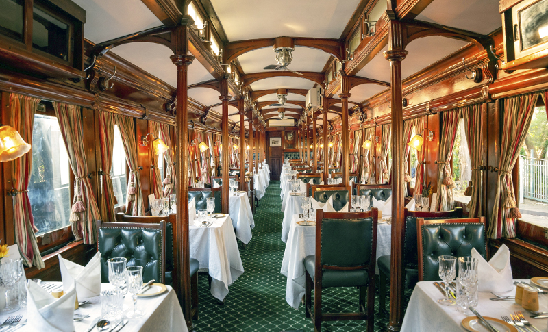 Vagón comedor del Bushtracks Express, con paneles de madera, asientos verdes y mesas preparadas con manteles blancos y copas.