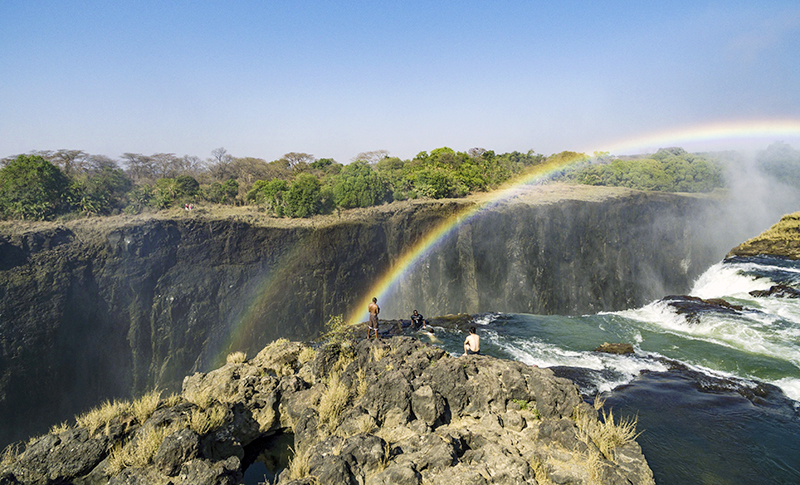 Visitantes sobre las rocas junto al salto de las Cataratas Victoria, en la zona de Livingstone Island, con un arcoíris entre la bruma.