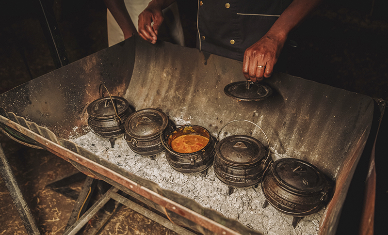Cocinero destapando una olla de potjie entre varias ollas de hierro sobre las brasas en The Eatery.
