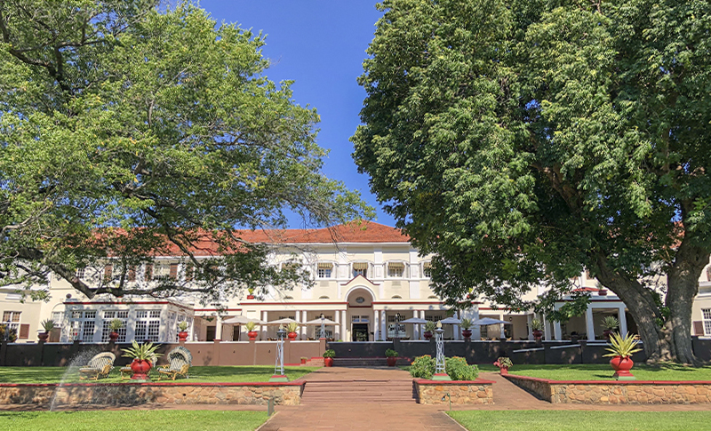 Fachada del Victoria Falls Hotel vista desde sus jardines, con grandes árboles y un camino central hacia la terraza.