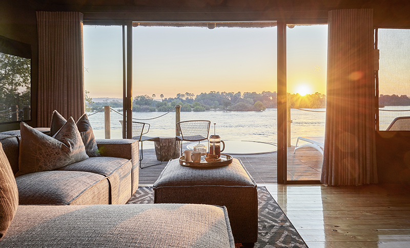 Salón de una suite de Victoria Falls River Lodge, con sofás y puertas de cristal abiertas a una terraza frente al Zambeze.
